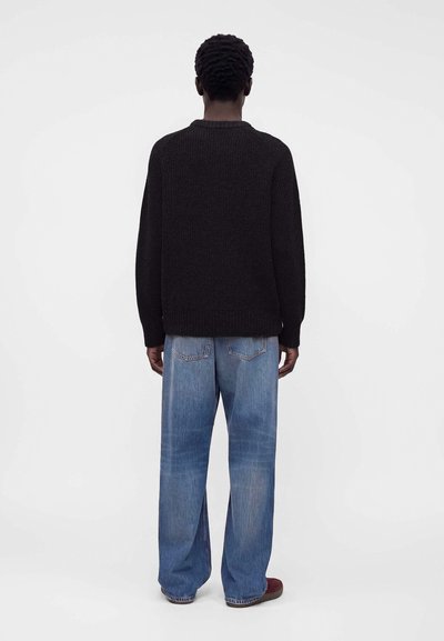 Homme debout, dos à la caméra, portant un pull noir, un jean bleu ample et des chaussures marron sur un fond clair uni.