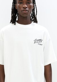 T-shirt overdimensionato bianco in cotone. Presenta un logo grafico nero sull'area sinistra del petto. Maniche corte e scollatura rotonda.
