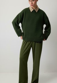 Pulover tricotat verde cu un guler bej, asortat cu pantaloni largi de culoare verde măsliniu. Materialul pare moale și texturat.