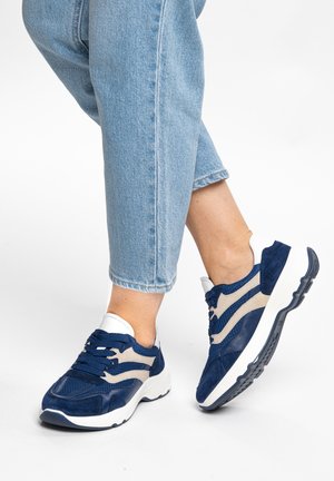 Person wearing light blue jeans and navy blue and white sneakers, standing with one foot slightly raised.