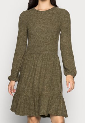 Kobieta w długiej sukience o długości do kolan w oliwkowym kolorze, z długim rękawem, z marszczonym paskiem w talii i falbanką na dole, stojąca na tle jednolitego tła.
