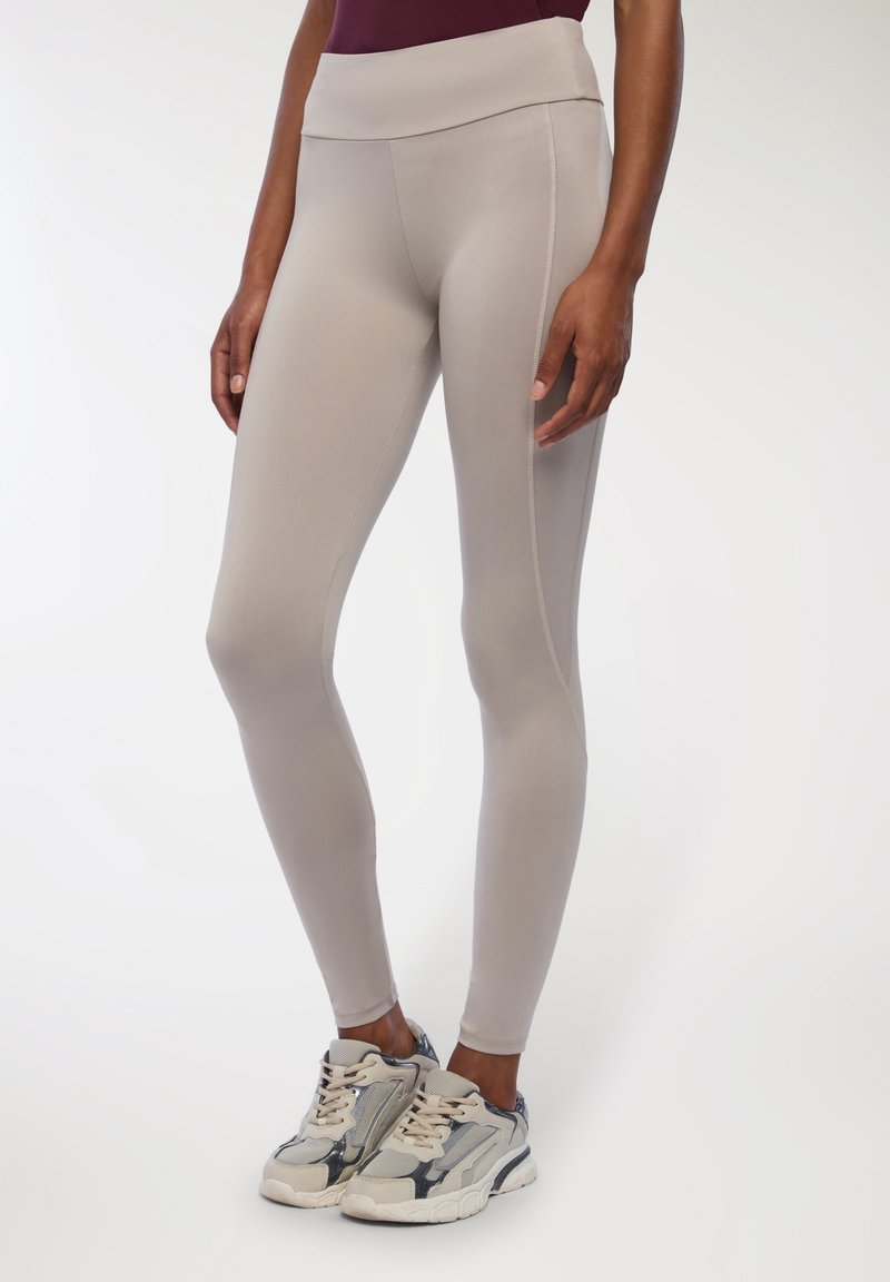 Beżowe legginsy z wysokim stanem, gładką fakturą i detalami na bocznych szwach, połączone z jasnoszarymi sneakersami z akcentami i sznurówkami.