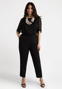 Top nero semi-trasparente con maniche corte abbinato a pantaloni neri a sigaretta. Una collana dichiarativa circolare bianca aggiunge contrasto. Tacchi neri con cinghie.