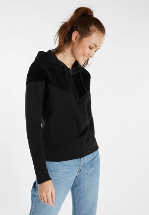 Zwarte hoodie met een textuur van ribfluweel over de schouders, met een rits aan de voorkant en geribbelde manchetten. Gekleed met lichtblauwe spijkerbroek.