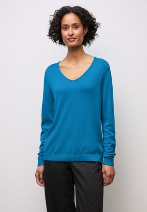 Pull en V turquoise en tricot léger et lisse. Les manches longues sont dotées de poignets côtelés ; l'ourlet est également côtelé. Associé à un pantalon sombre.