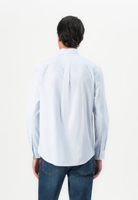 Mann med mørkt hår iført en lys blåmønstret skjorte med lange ermer og blå jeans, sett fra bakgrunnen mot en hvit bakgrunn.
