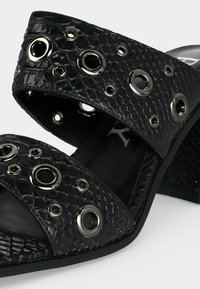 Μαύρο υφασμάτινο πέδιλο με τακούνι, διπλές λουρίδες με μεγάλα ασημένια μεταλλικά ρούχα και λεπτομέρεια με εξαγωνικό μοτίβο.