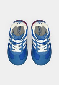 Zili sporta apavi ar baltiem svītrām, sintētiska augšdaļa, noapaļota purngala un baltas šnores. Sarkanīgi akcenti uz zoles; zīmola interjers.