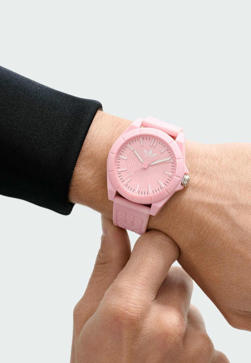 Montre en silicone rose avec un cadran texturé, des marqueurs d'heure simples et une couronne métallique. Portée au poignet avec une chemise à manches longues noire.