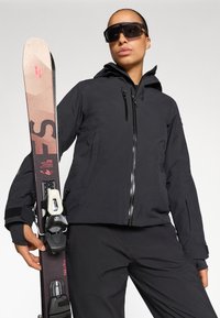 Schwarze wetterfeste Jacke mit Reißverschluss, strukturiertem Stoff und verstellbare Kapuze. Hält Skier mit metallischer Bindung und beigem Ende. Schwarze Sonnenbrille.