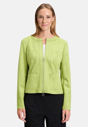 Veste en suède verte avec un col rond, fermeture éclair à l'avant, deux poches sur la poitrine et une coupe cintrée, portée sur un pantalon noir.