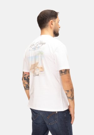 Uomo con capelli corti e scuri e tatuaggi sulle braccia, indossa una maglietta bianca con una scena di spiaggia e testo sul retro, abbinata a jeans blu.