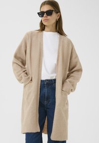 Moteris, vilkinti juodus katės akies akiniai, smėlio spalvos ilgas kardiganas, balta marškinėliai ir mėlyni aukšto juosmens džinsai, stovi su rankomis kišenėse.