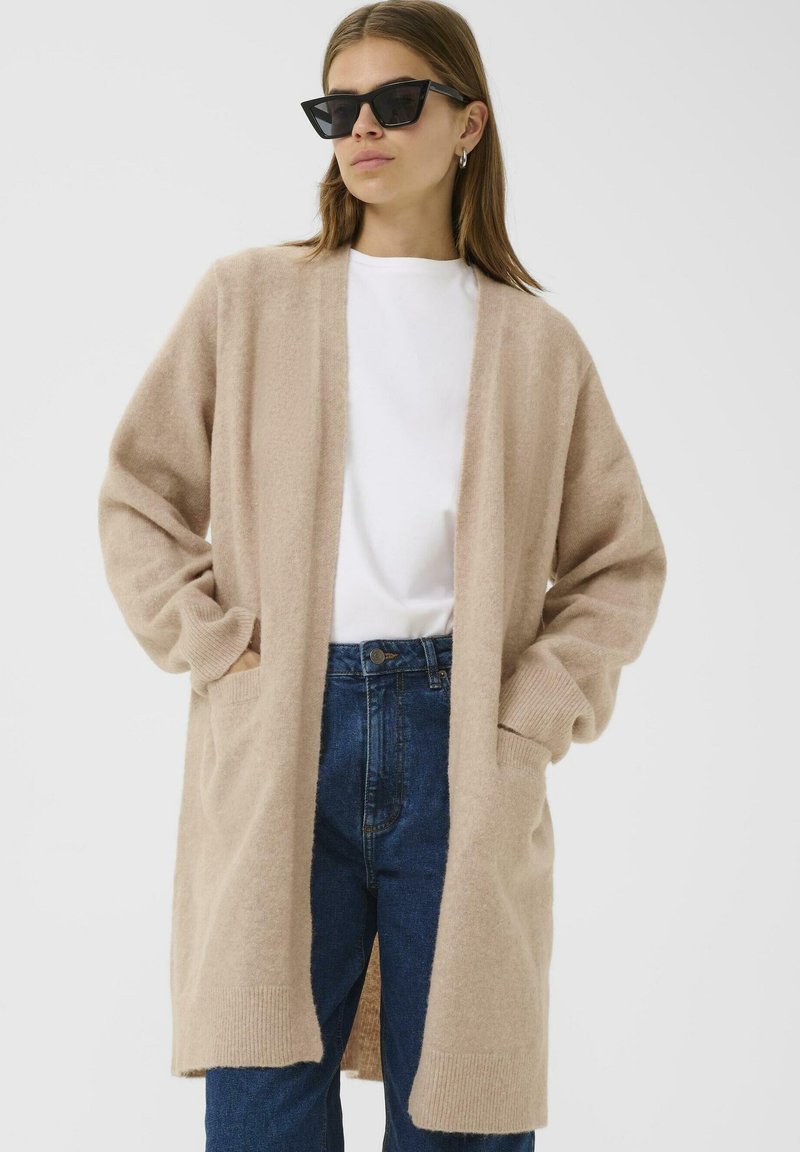 Moteris, vilkinti juodus katės akies akiniai, smėlio spalvos ilgas kardiganas, balta marškinėliai ir mėlyni aukšto juosmens džinsai, stovi su rankomis kišenėse.