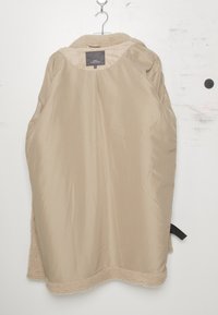 Beige isolerad jacka med en slät yttermantel och ett mjukt, strukturerat innerfoder. Har en rundad krage och en sidrem.