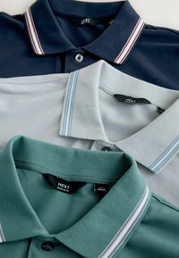 Trois polos empilés : bleu marine foncé avec des rayures rouges et blanches, gris clair avec des rayures bleues, et vert, tous avec un col à boutons.