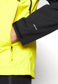Giacca impermeabile gialla e nera con tasca con zip e chiusura con velcro sui polsini; presenta il logo "DRY VENT" sulla manica.