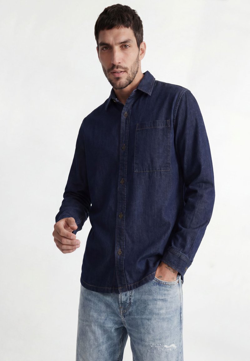 Mann trägt ein dunkelblaues, langärmliges Hemd mit Knöpfen und hellblaue Jeans, steht mit einer Hand in der Tasche vor einfarbigem Hintergrund.