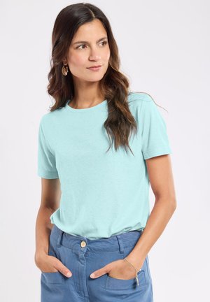Femme aux longs cheveux bruns portant un t-shirt bleu clair et un pantalon bleu, mains dans les poches, face légèrement tournée vers la gauche, fond neutre.