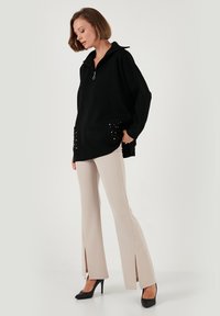 Un sweat à zip noir avec de longues manches et deux poches ornées. Un pantalon beige évasé avec des fentes latérales. Modèle portant des talons noirs.