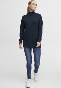 Dunkelblauer Rollkragenpullover mit gerippten Bündchen und Saum, kombiniert mit dunklen Jeans und weißen Sneakern. Der Pullover hat eine lockere Passform.