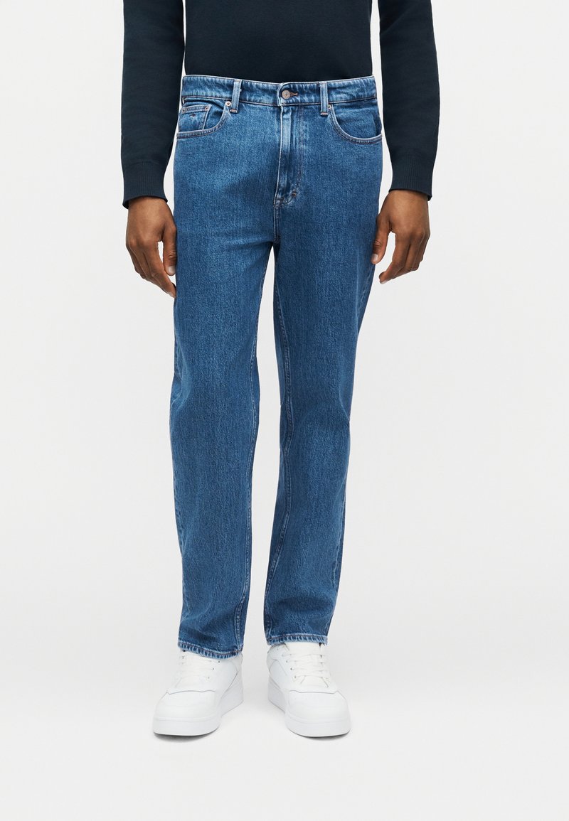 Άνδρας που φοράει μπλε τζιν παντελόνι σε ίσια γραμμή, λευκά αθλητικά παπούτσια και σκούρο πουκάμισο με μακρύ μανίκι, στέκεται μπροστά από απλό φόντο.