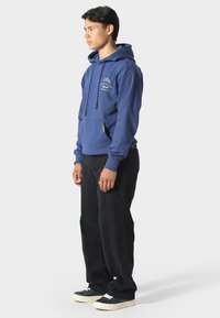 Felpa blu navy con tasca a marsupio, cordini regolabili e dettaglio ricamato sul fronte, abbinata a pantaloni neri e sneaker.