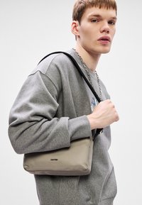 Šedá crossbody taška z látky s hladkým povrchem, zipovým uzavíráním a černým nastavitelným popruhem. Na přední straně je decentní logo Calvin Klein.