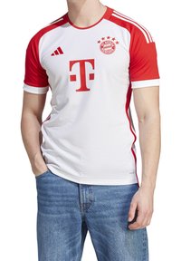 Bayern München jalkapallopaita, valkoinen ja punainen, lyhyillä hihoilla, jossa on suuri 'T' logo, seuran vaakuna ja kolme raitaa olkapäillä.