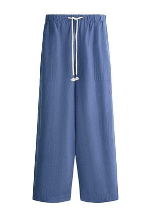 Kobieta idzie ubrana w niebiesko-białą koszulę w paski z długim rękawem, luźne niebieskie spodnie, czarne sandały, niosąc dużą ciemnoniebieską torbę typu tote.