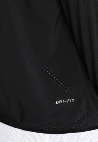 Černé sportovní tričko Dri-Fit s perforovanou texturou, s bílým logem na spodní straně. Lehký, prodyšný design.