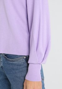 Fioletowy sweter z przędzy z teksturowanym materiałem, długimi rękawami w kształcie balonu i ściągaczami. Noszony z niebieskimi dżinsami.