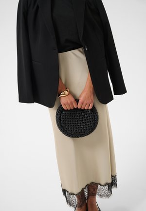 Donna con gonna beige con orlo in pizzo nero, blazer nero, braccialetto dorato, che tiene una piccola borsa nera intrecciata davanti a uno sfondo semplice.