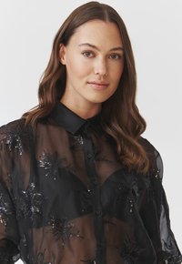 Bluză neagră transparentă cu paiete aranjate în motive florale, având un închidere cu nasturi și un guler clasic. Textură netedă și un design ușor.