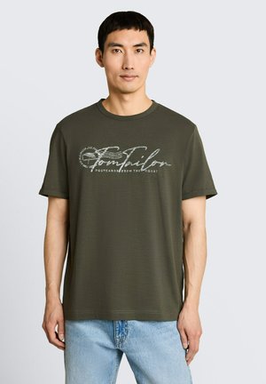 Jonge man met kort zwart haar die een olijfgroen T-shirt met de tekst "Tom Tailor" en lichtblauwe spijkerbroek draagt, staand tegen een effen achtergrond.