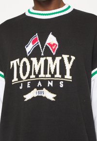 Mustassa neulepaidassa on valkoiset "TOMMY JEANS" -kirjaimet ja brodeeratut liput ylhäällä. Ribbaatut vihreä ja valkoinen kaulus. Pehmeä rakenne.