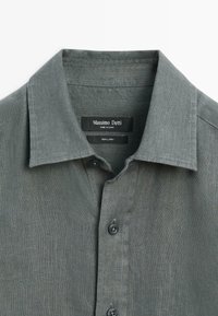 Chemise en lin grise avec col pointu et boutons noirs, présentant l'étiquette de la marque "Massimo Dutti" à l'intérieur du col.