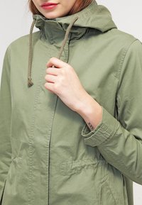 Olivgrön huva jacka med dragkedja, snörningsdetaljer och två framfickor med en mjuk, slät textur.