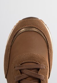 Baskets marron avec détails en daim et tissu, bout rond, lacets marron assortis sur un fond clair uni.