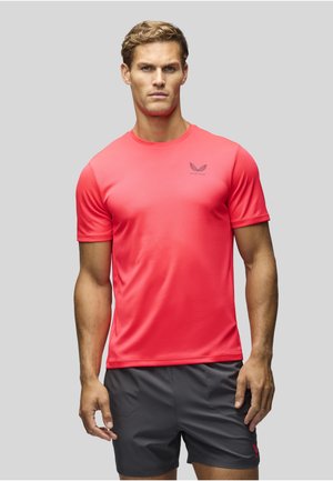 Homme aux cheveux blonds courts portant un t-shirt de sport rouge vif et un short gris foncé, debout devant un fond uni gris clair.