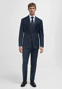 Blazer bleu marine foncé avec un pantalon assorti. Fabriqué en tissu lisse, à une seule rangée de boutons, et complété par une chemise blanche et une cravate.