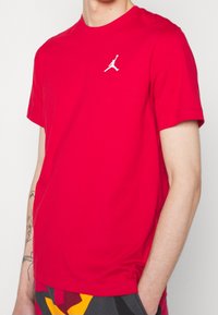 Hombre que lleva una camiseta de manga corta roja brillante con un pequeño logo blanco de Jumpman en el pecho izquierdo y pantalones de patrón colorido.