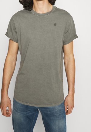 Homme portant un t-shirt vert olive à manches retroussées et un jean bleu, debout devant un fond blanc uni, torse visible du menton jusqu'aux hanches.