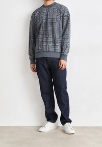 Suéter gris con estampado repetido de "HUGO", puños y dobladillo acanalados. Combinado con jeans azul oscuro y zapatillas blancas. Estilo simple y casual.