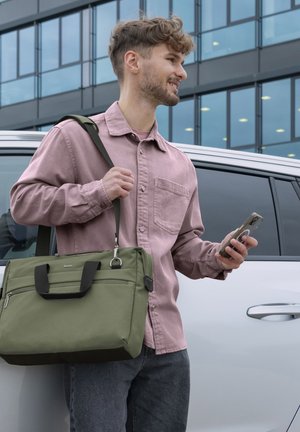 Junger Mann in einem pinken Hemd, der ein Smartphone hält und eine olivgrüne Umhängetasche trägt, steht neben einem weißen Auto mit einem Hintergrund aus Glasgebäuden.