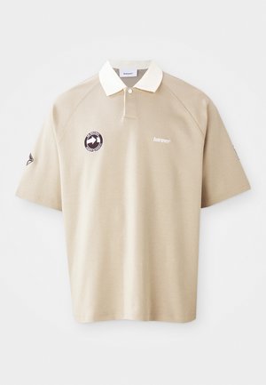 Vaalean beige lyhythihainen polo-paita, jossa on valkoinen kaulus, nappilista, brändilogo rinnassa ja pyöreä pata vasemmalla puolella.