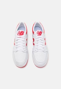 Vita sportskor i vitt med röda accenter, som har en perforerad tårbox, snören och en stor "N"-logotyp på sidan. Släta läder- och mesh-material.