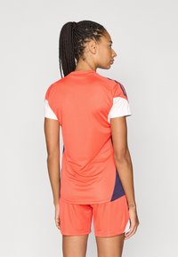 Αθλητική στολή που περιλαμβάνει φωτεινή κοραλί μπλούζα με λευκές και ναυτικές λεπτομέρειες και ταιριαστά κοραλί σορτς, κατασκευασμένα από ελαφρύ, διαπνέον υλικό.