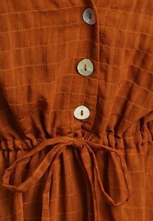 Rust-colored dress with a checkered pattern, featuring four round, light-colored buttons and a tied waist. Textured fabric with a soft finish.