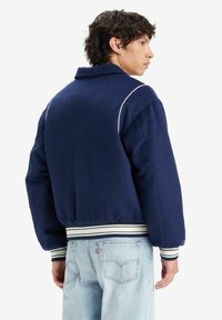 Navy blue padded jacket with a wide collar, white piping accents, and striped cuffs. Paired with light blue denim jeans.